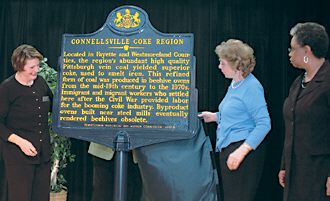 (From left) Pamela Seighman, Elaine DeFrank and Karen James, administrative officer for Bureau of Archive and History of the Pennsylvania Historical and Museum Commission, unveil the Connellsville Coke Region Historical Marker at Swimmer Hall at Penn State Fayette, The Eberly Campus. Hidden behind the sign is Dr. Evelyn Hovanec, associate professor of English and American Studies, emerita director of the Coal and Coke Heritage Center. Ed Cope/Herald-Standard